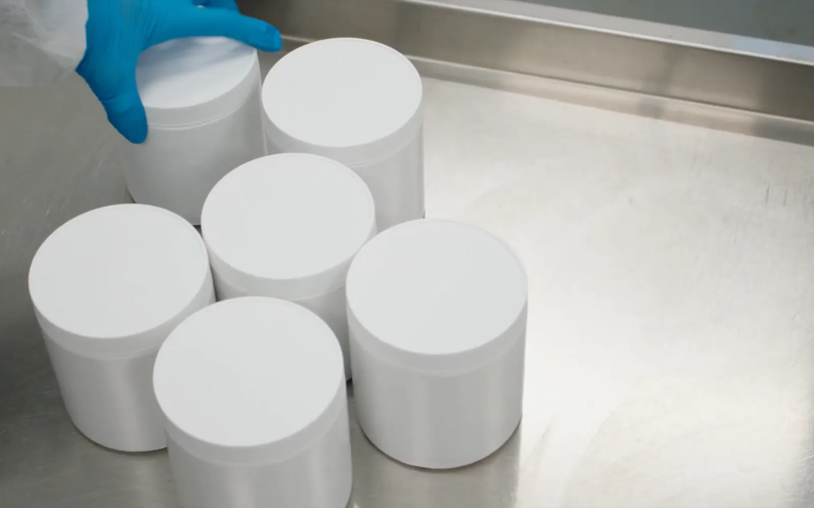 Six plain white supplement tubs on the stainless steel bench, a gloved hand setting one down, ready for a label